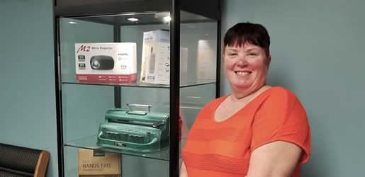 A smiling woman next to a glass display case with lights showing off three shelves of equipment including a wireless projecor, pocket talker, braille embosser, and a box that says "hands free". 
