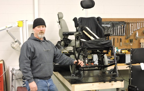 A man stands next to a wheel chair on a table with tools mounted behind. 