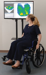 A woman seated in a wheelchair looks up at a digital pressure mapping image on a display behind her.