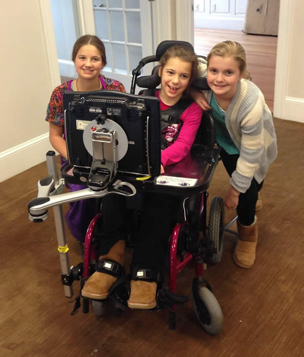 Smiling girl seated in wheelchair faces mounted monitor with her two sisters crouched smiling by her sides.
