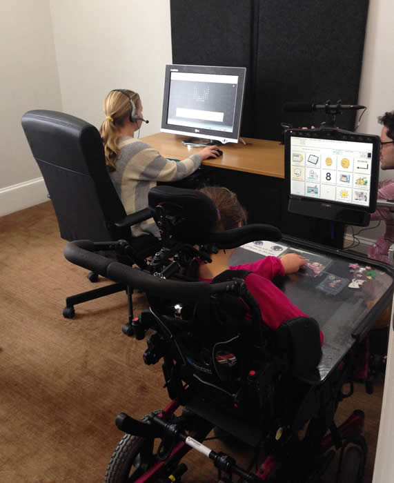 Two children seated in front of monitors. One wears a headset with a mic. The other is in a powerchair wth headrest and communication display.