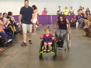Wheelchair hip hop instructor chasing after a gleeful 2-year-old boy in his wheelchair with his father smiling behind and the audience clapping. 