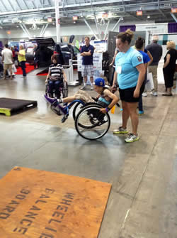 A boy attempts popping a wheelie in a manual chair with the help of a demo attendee. 