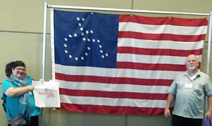 A man and a woman stand smiling in front of a large U S flag with stars aligned to shape a wheelchair symbol. The woman holds an Abilities Expo sign.