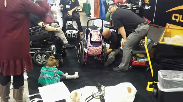 People gathered at an exhibition booth, some in wheelchairs, a small girl wears a leg brace seated on the floor. Two vendors bend over her manual wheelchair for a repair. Service dog in foreground.