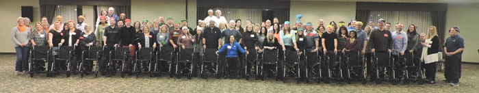 Group shot of dozens of adults standing and smiling behind a line of 16 wheelchairs. One woman seated in the middle. 