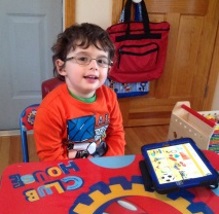 Young boy seated and smiling at the camera, his tablet on the table in front of him.